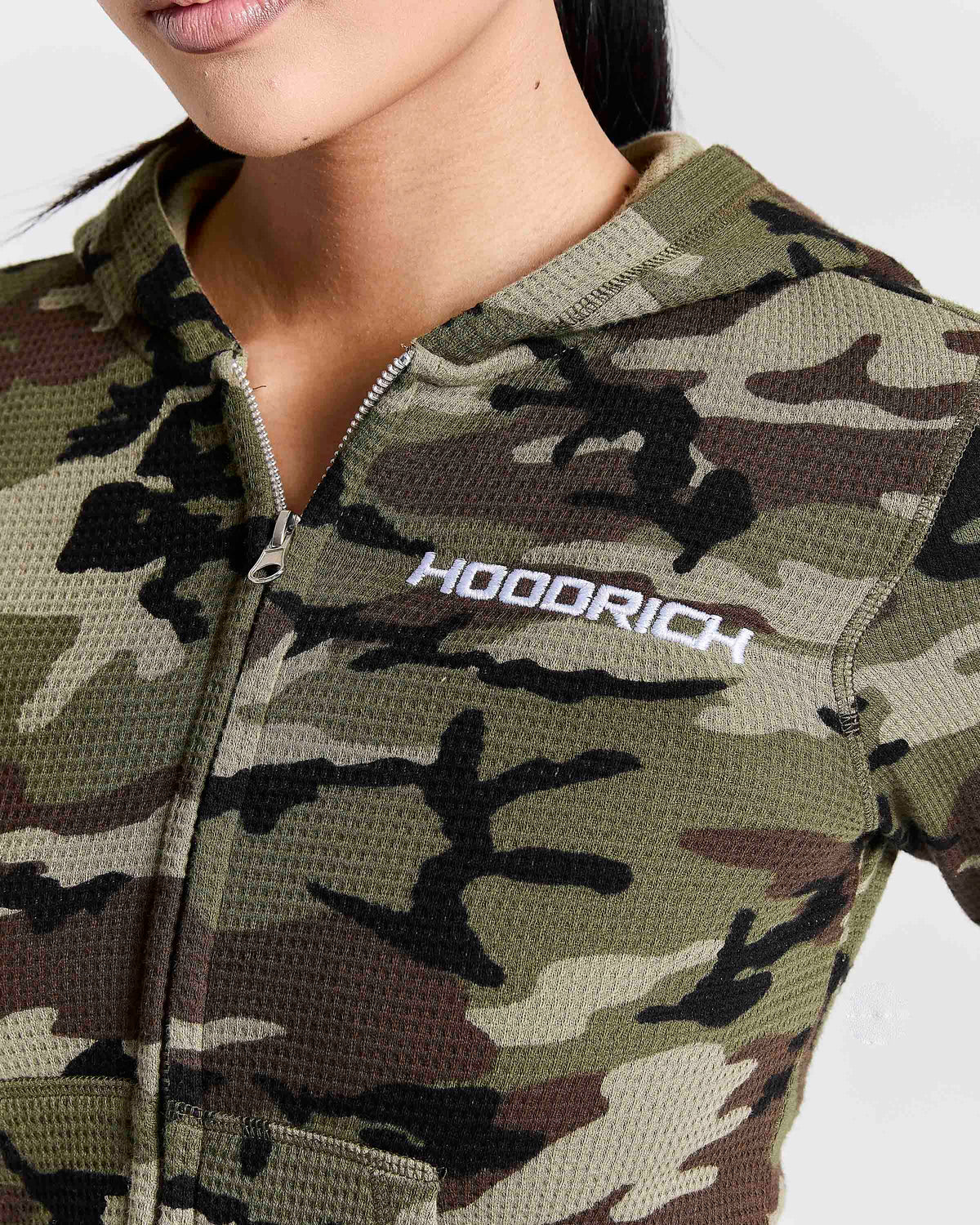 Close-up of a woman wearing a camouflage zip-up hoodie with the brand name 'HOODRICH' printed in white on the chest.