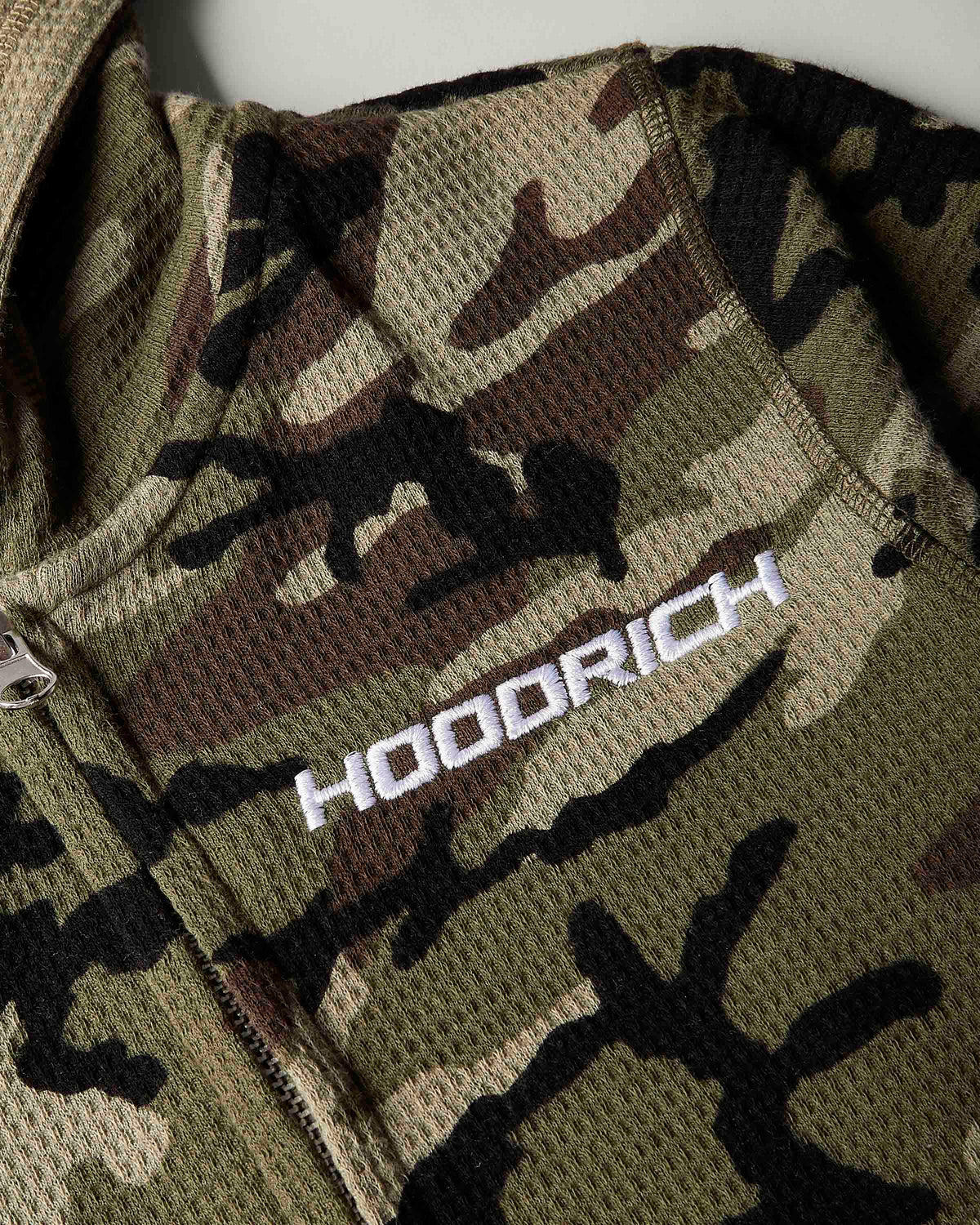 Close-up of a camouflage mesh zip jacket with a white embroidered 'HOODRICH' logo on the chest.