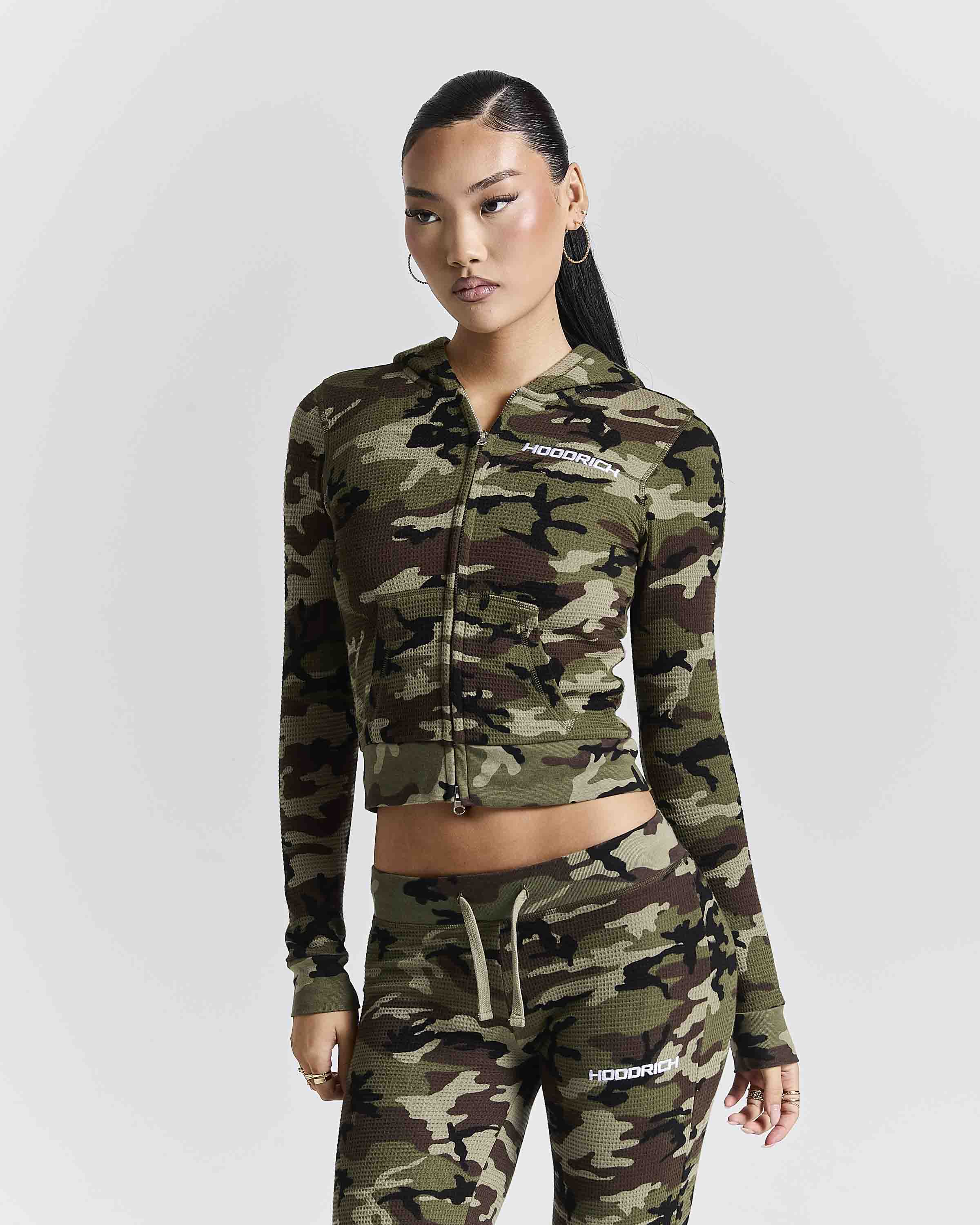 A model wearing a matching camouflage zip-up hoodie and jogger pants set with white HOODRICH branding, photographed against a light gray studio background.