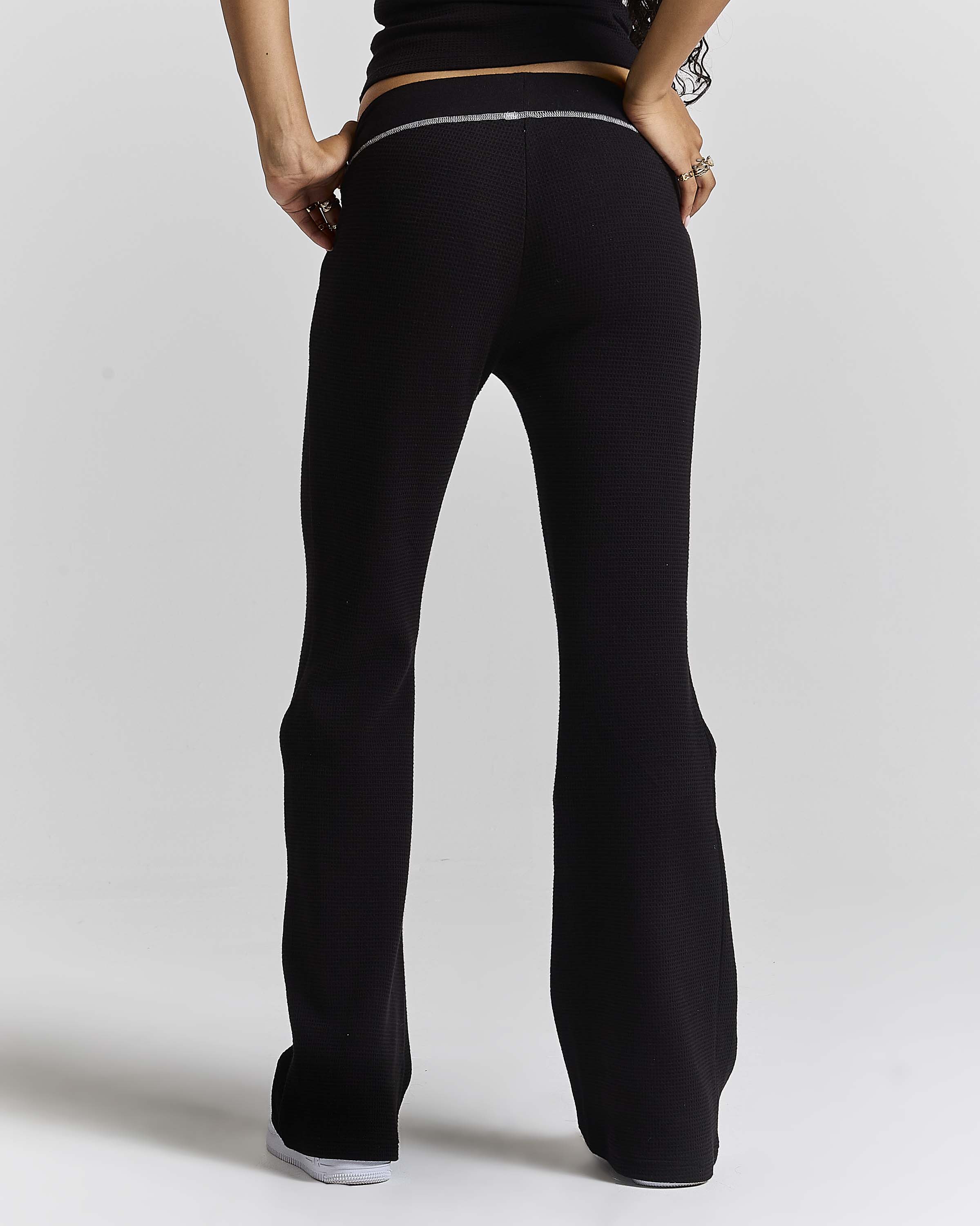 Rear view of a model wearing black flared pants with a thin silver metallic trim detail at the back waistband, set against a plain light gray studio background.