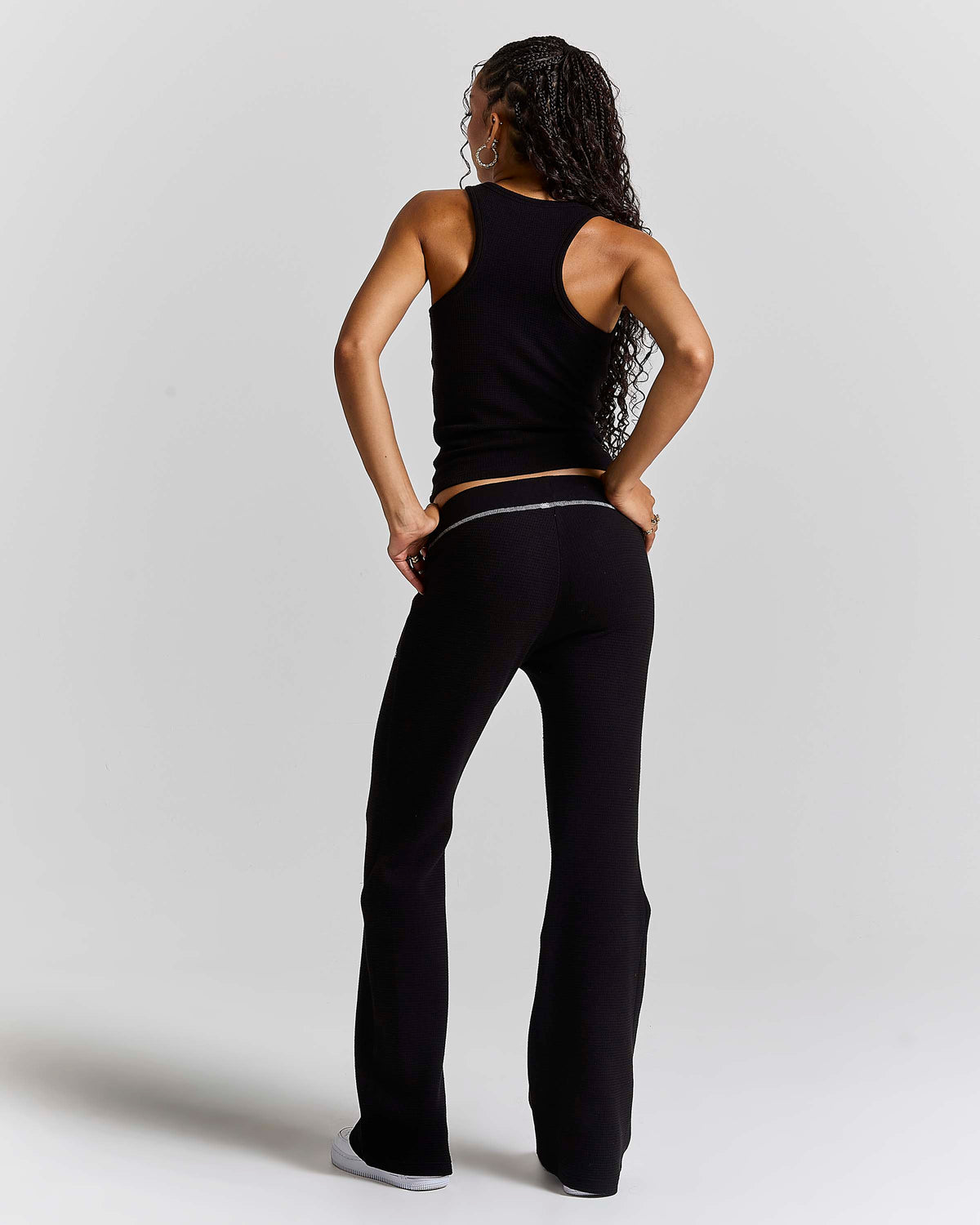 A woman shown from the back wearing a black ribbed tank top and matching black flared pants with a thin silver waistband, standing against a light gray studio background.