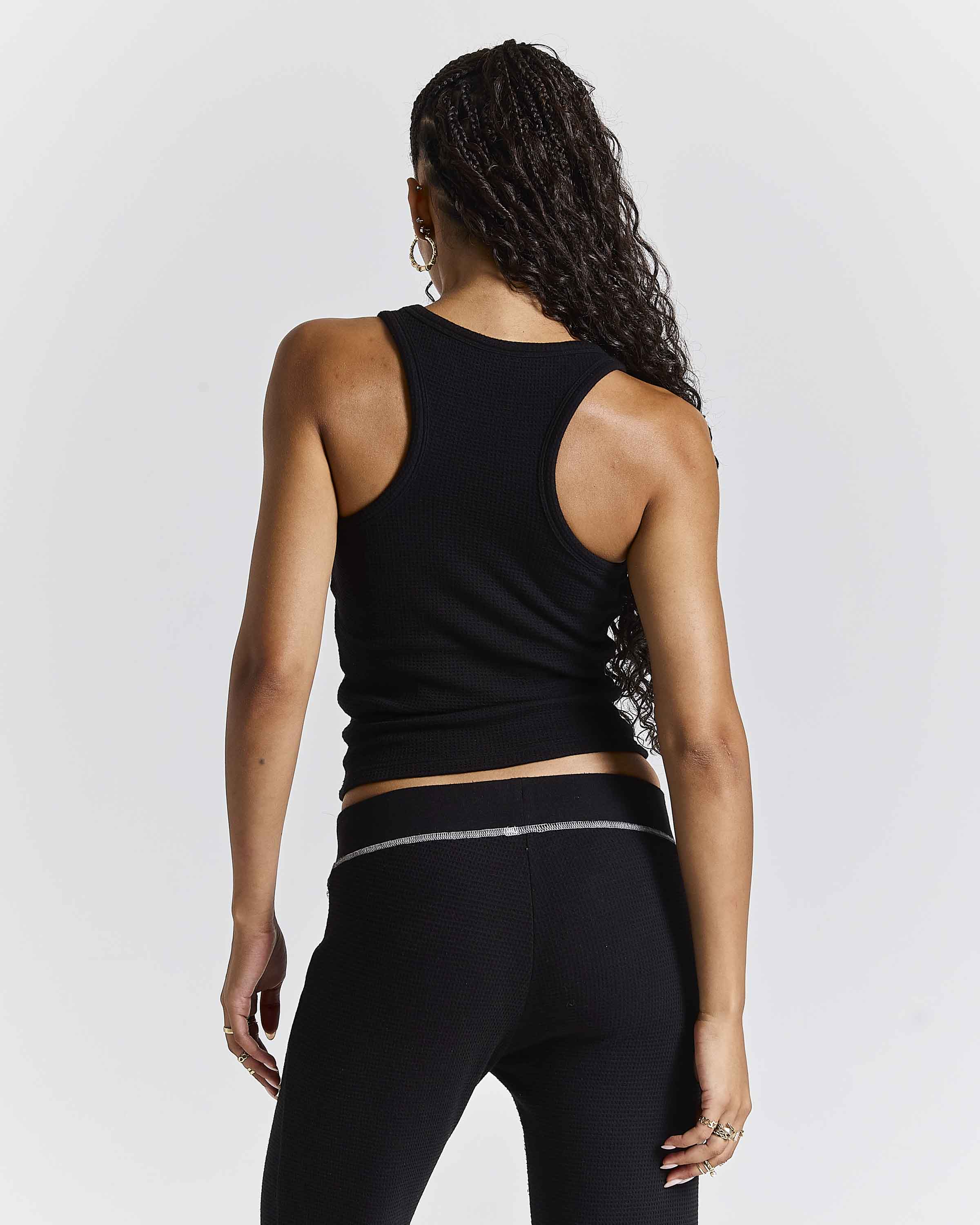 Rear view of a woman wearing a black ribbed athletic tank top and black leggings with a thin silver stripe detail on the waistband, set against a light gray studio background.