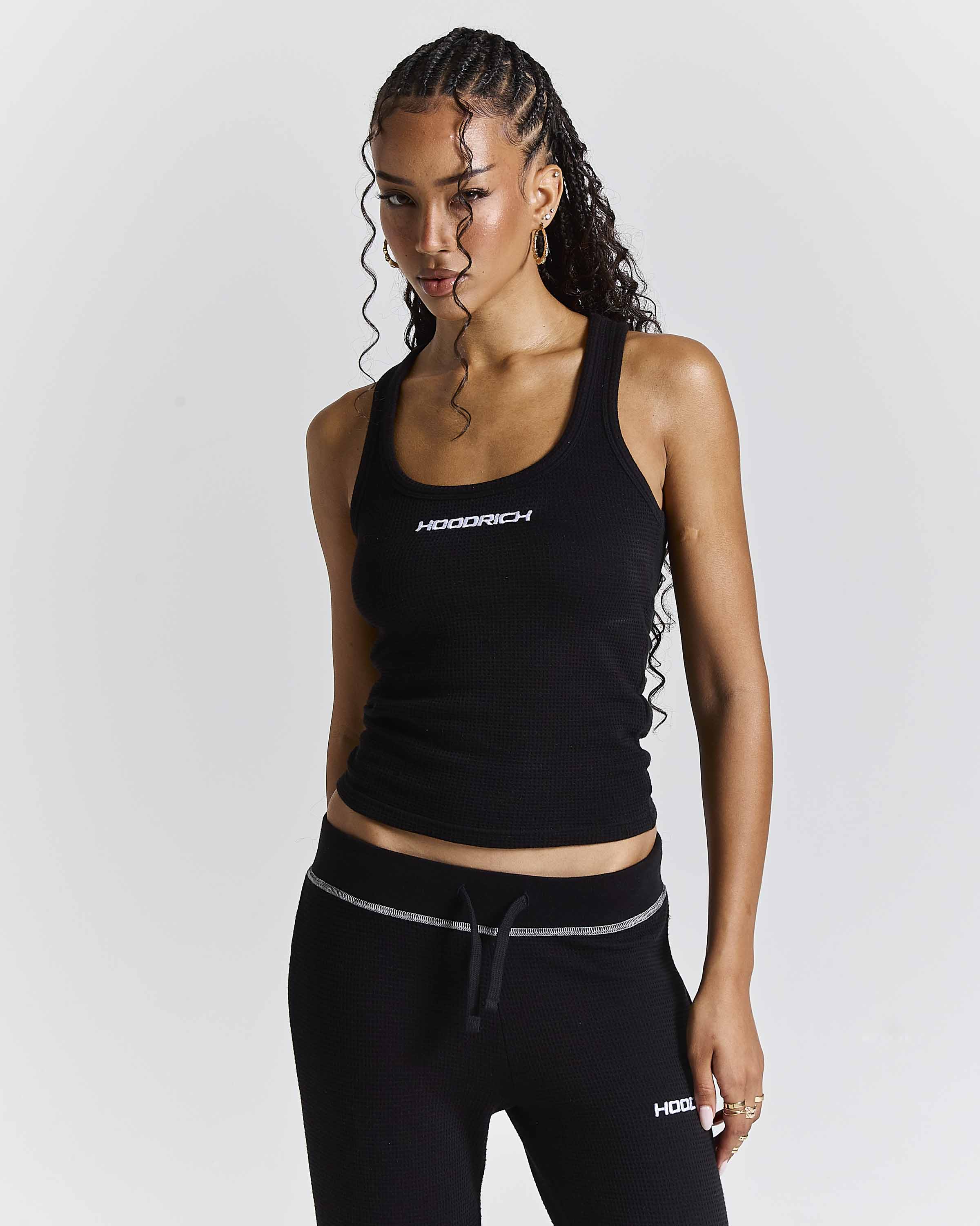 A woman modeling a black ribbed athletic tank top and matching black joggers with white accents, featuring the brand name 'HOODRICH' on the chest and upper thigh.