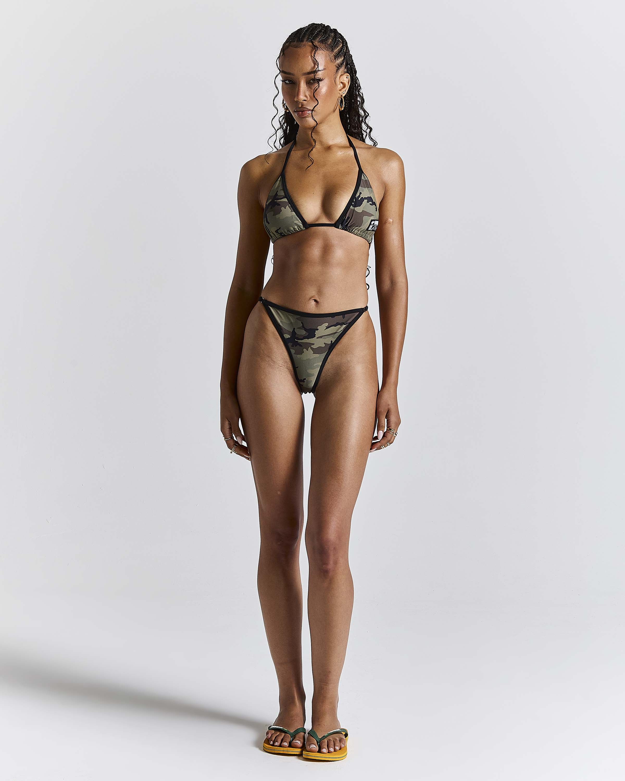 A woman modeling a two-piece bikini set with a green and black camouflage pattern, standing against a light gray studio background.