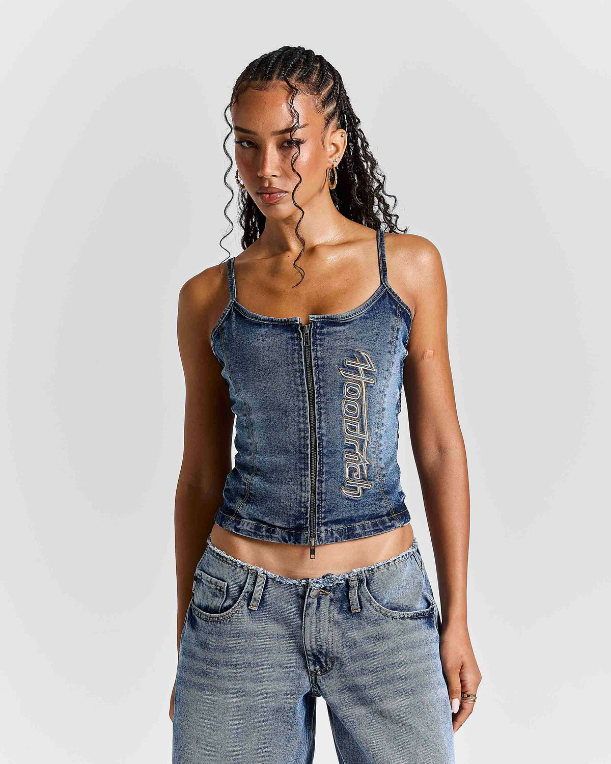 A woman modeling a cropped blue denim camisole top with a front zipper and vertical 'Hoodrich' text on the front, paired with light-wash jeans against a light gray studio background.