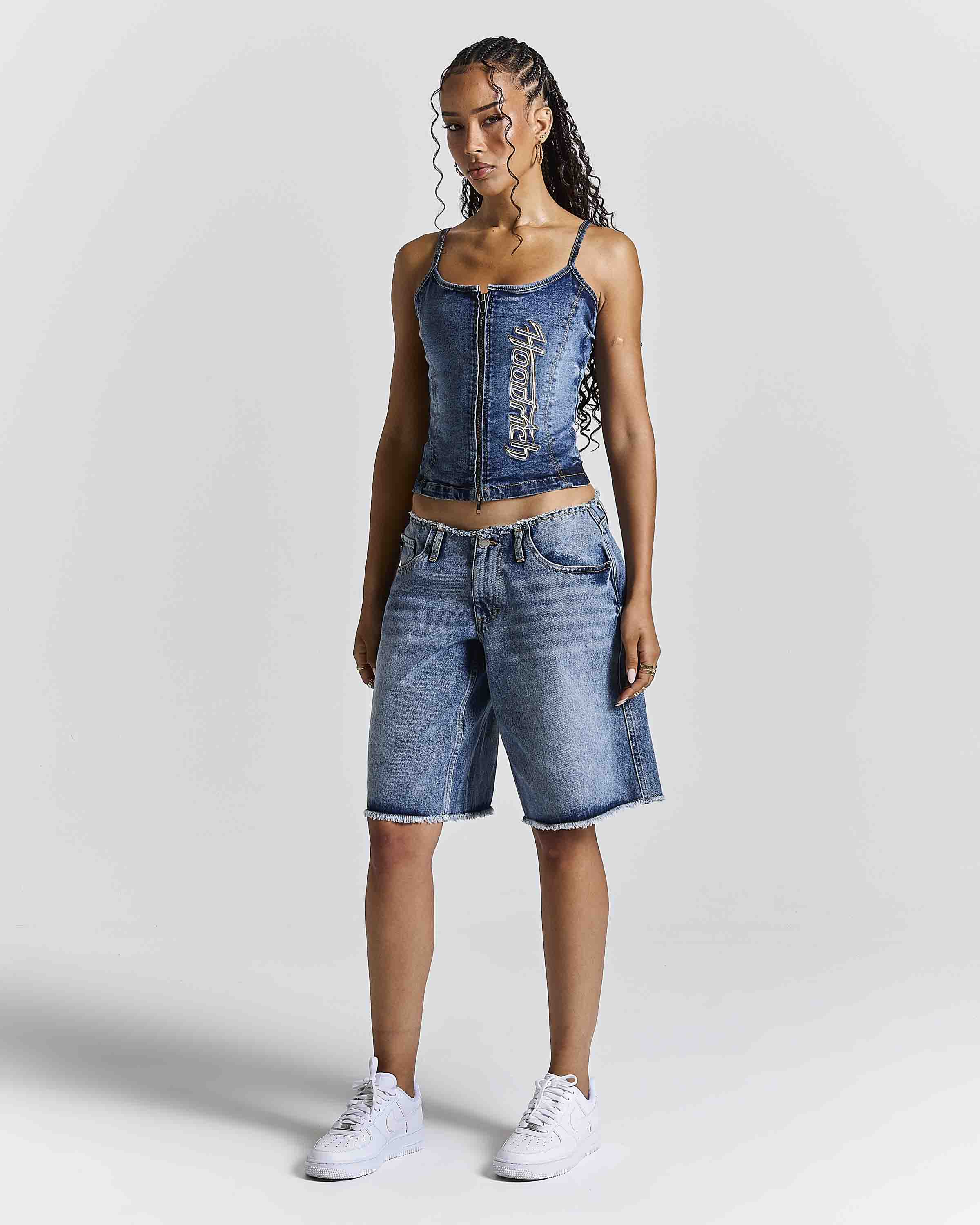 A woman modeling a matching denim set consisting of a blue denim camisole top with silver embellishments and blue denim shorts, paired with white sneakers, against a light gray studio background.