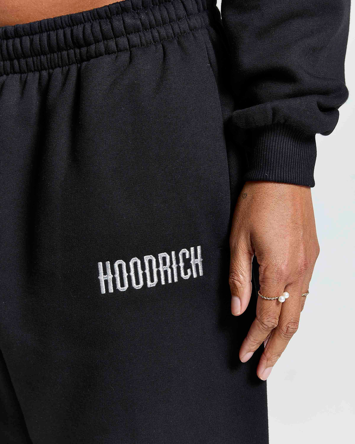 Close-up of a person wearing black sweatpants with the brand name 'HOODRICH' embroidered in white on the thigh, paired with a black long-sleeve top.