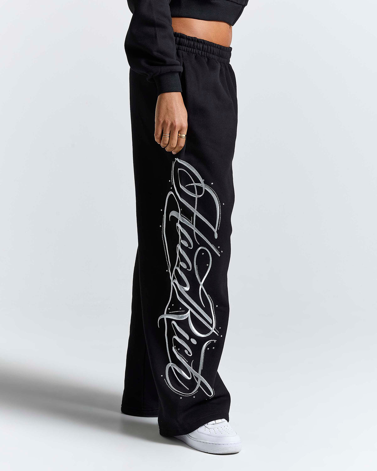 A person wearing black wide-leg sweatpants with a large white cursive graphic print down the left leg, paired with a black cropped top and white sneakers, photographed against a light gray studio background.