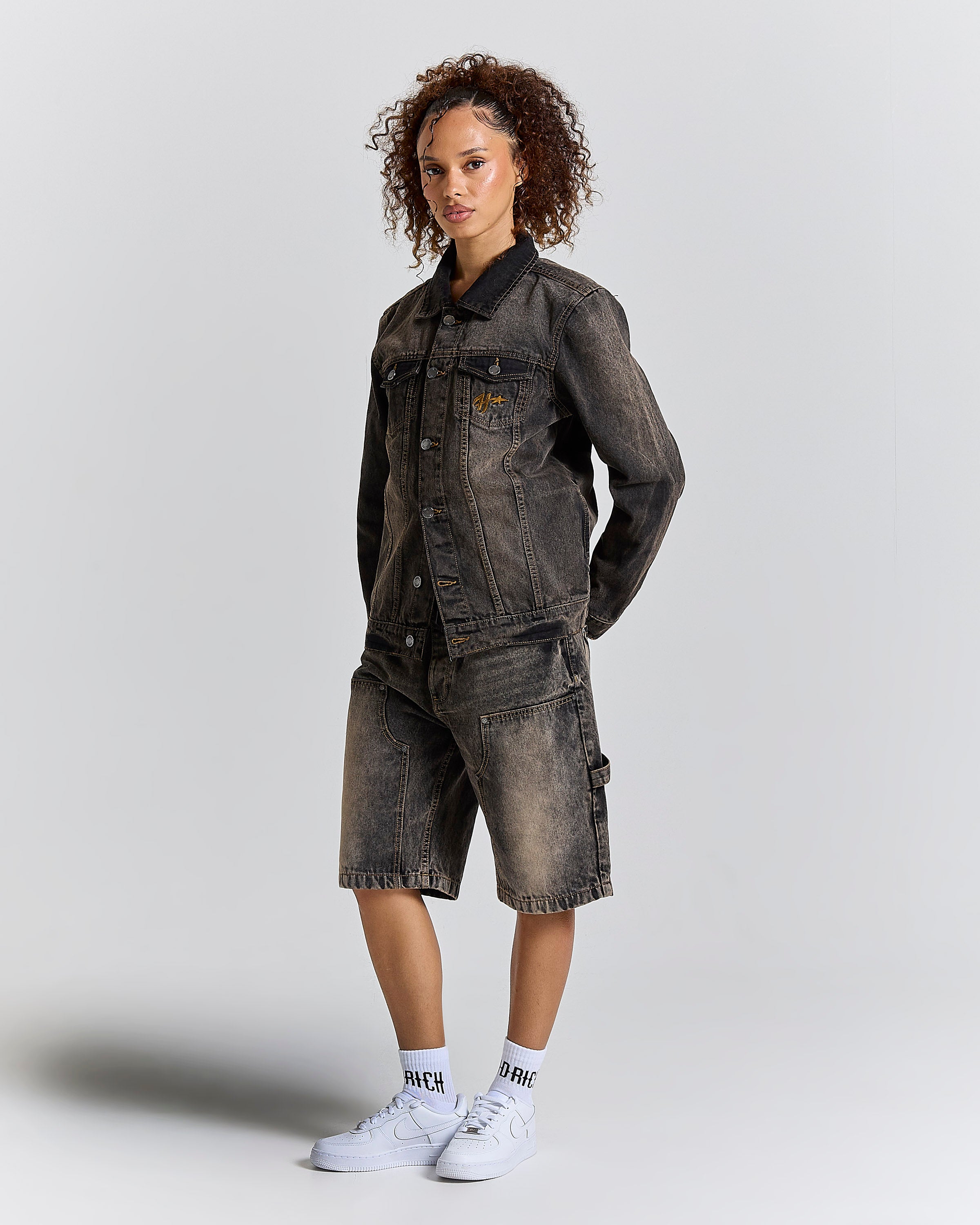 A model wearing a dark gray acid-washed denim jacket and matching denim shorts, styled with white crew socks and white sneakers, against a light gray studio background.
