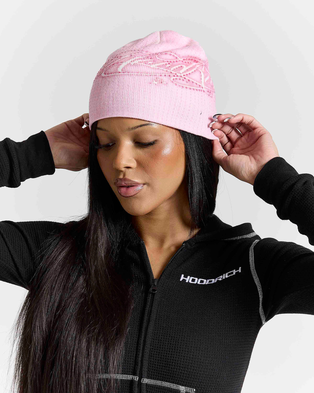 A woman wearing a light pink knit beanie with a white embroidered crown design, paired with a black zip-up hoodie featuring the brand name HOODRICH.