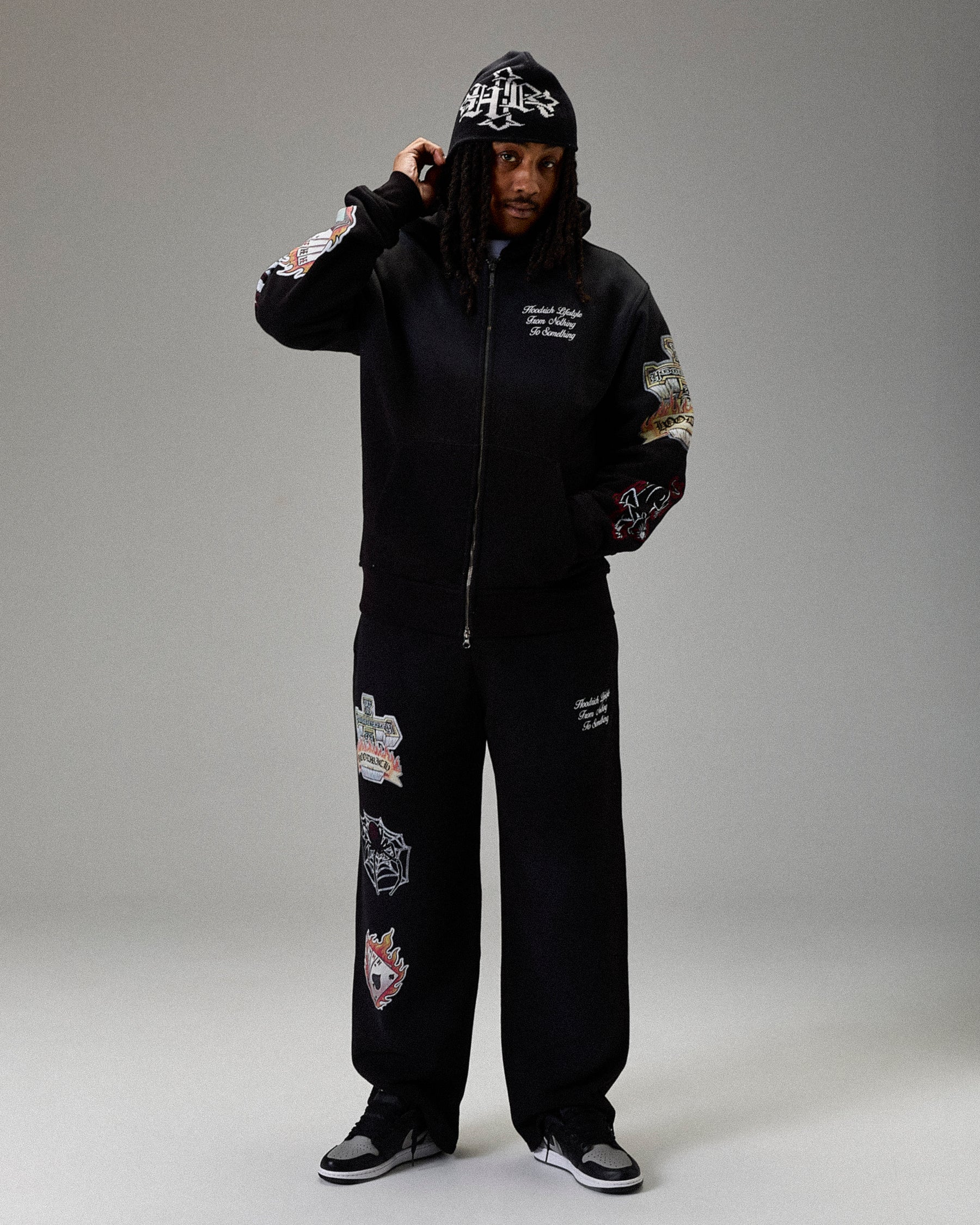 Person wearing a black hoodie and pants with patches on a gray background