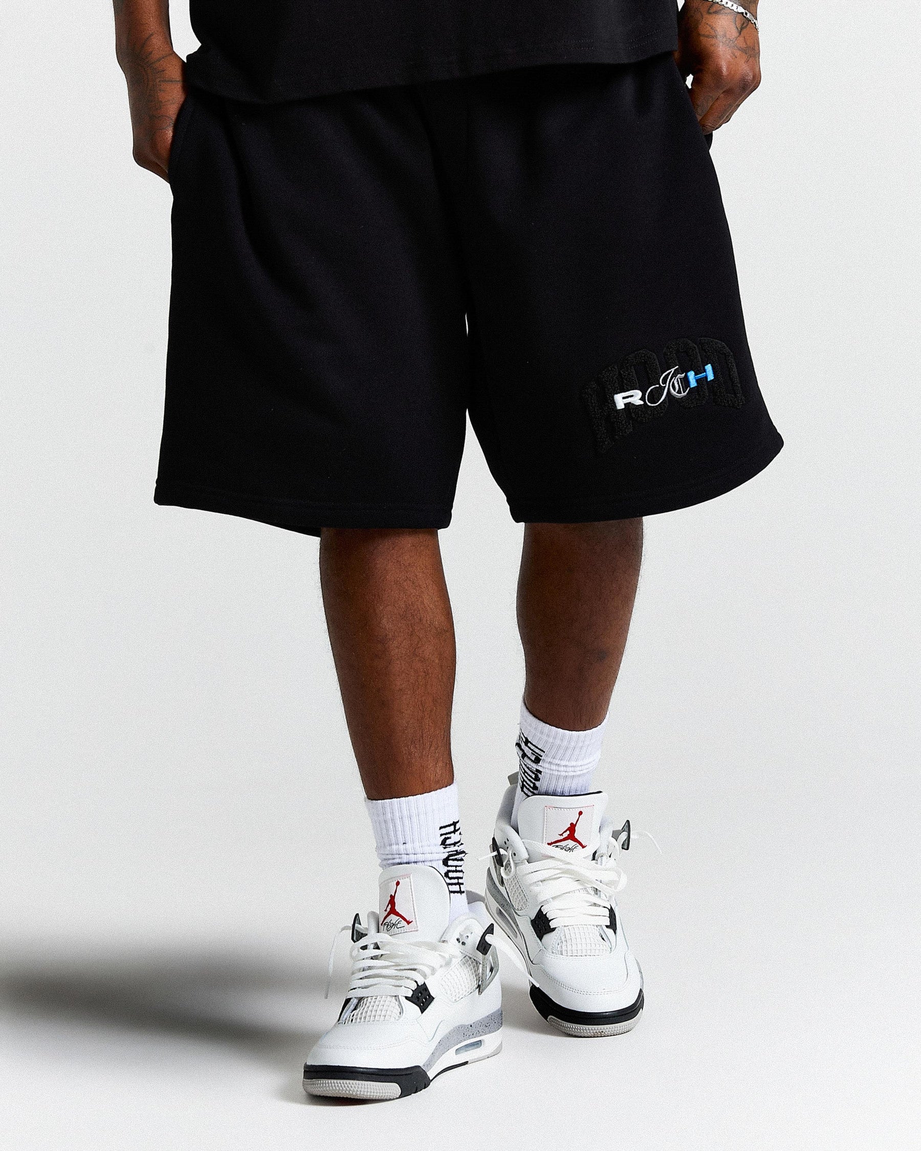 Person wearing black shorts with a logo and white sneakers on a light gray background