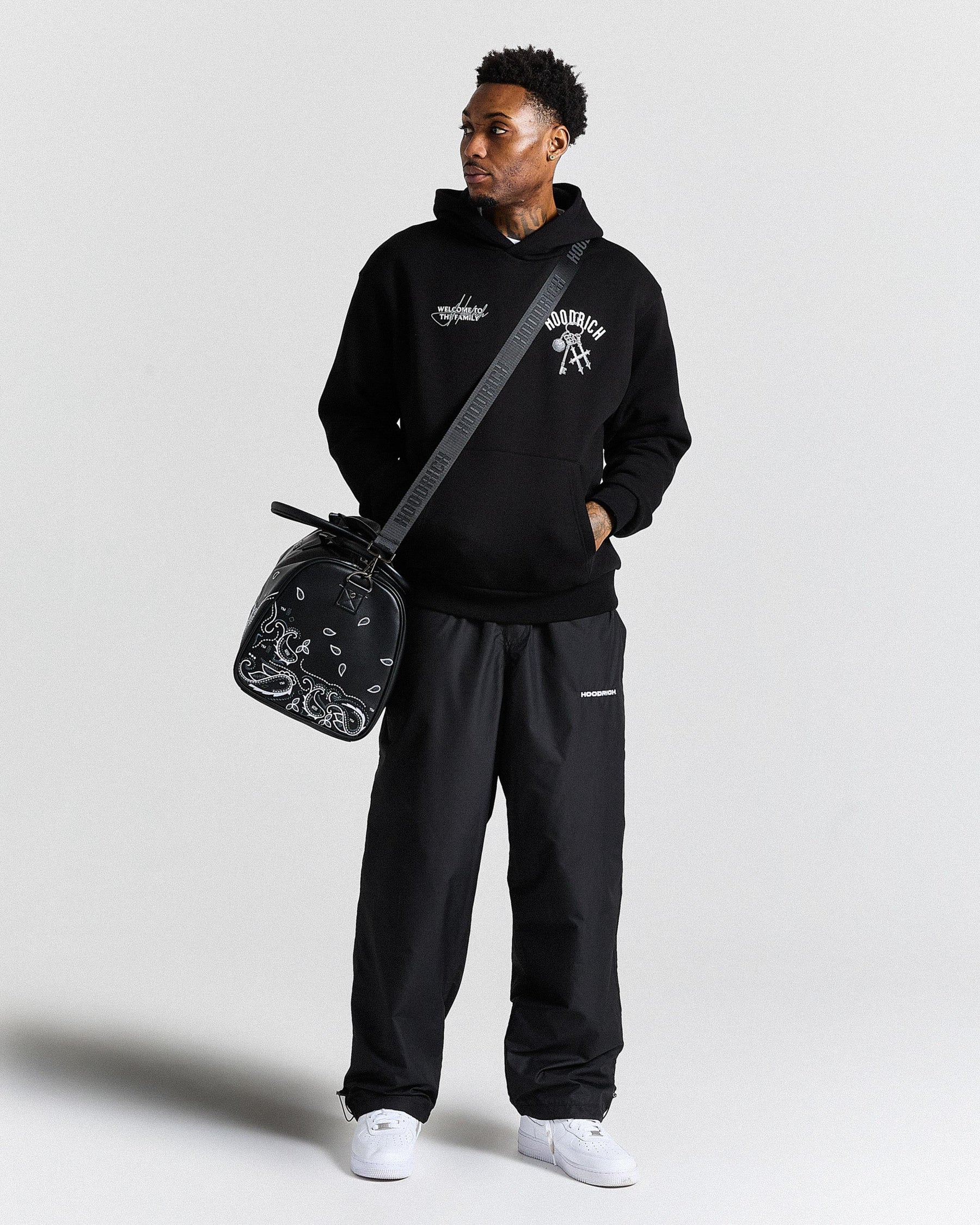 Person wearing a black hoodie and pants with a bag, standing against a plain background