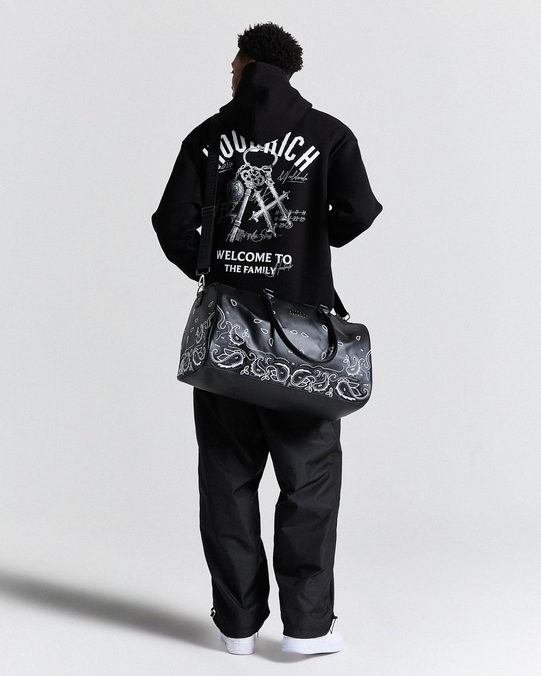 Person wearing a black hoodie with text and graphics, holding a black bag with white designs on a light gray background.