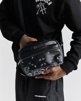 Person holding a black bag with white patterns, wearing a black hoodie and pants with 'Hoodrich' branding.