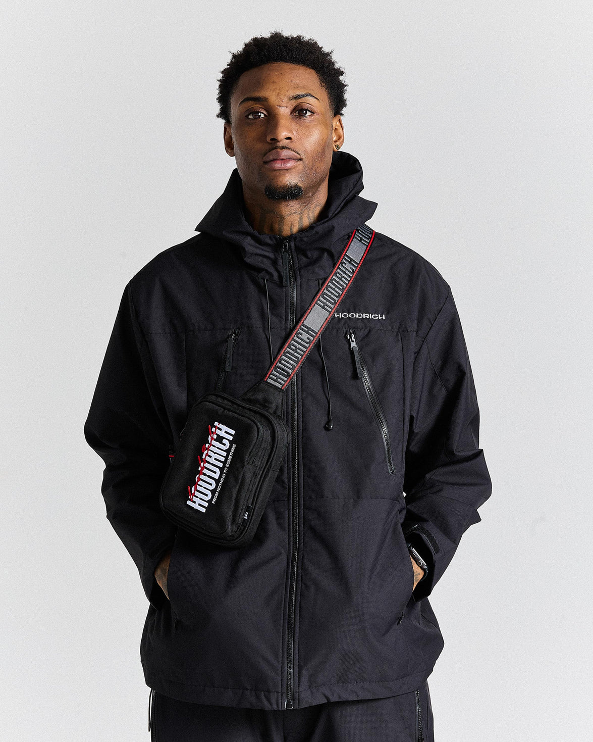 Man wearing a black jacket and a cross body bag with a visible 'Hoodrich' logo on a plain background