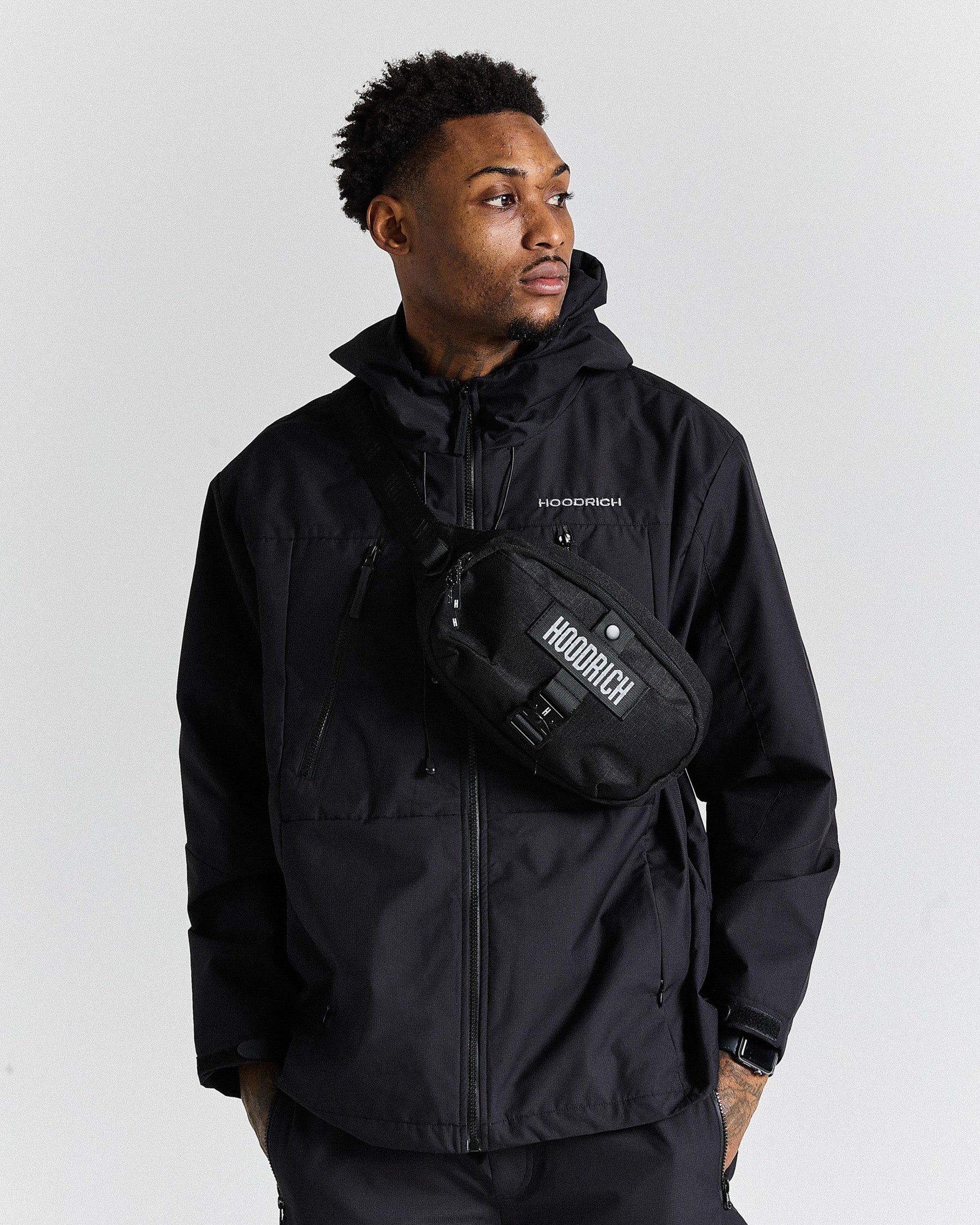Man wearing a black jacket with a hood and a matching waist bag, standing against a plain background.