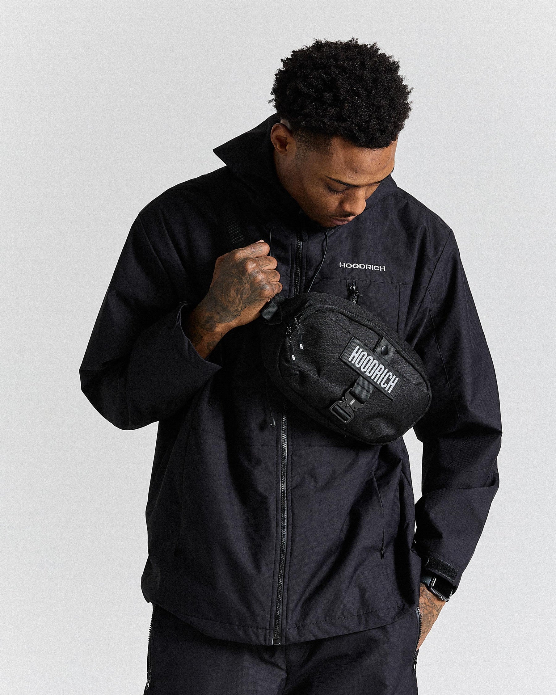 Person wearing a black jacket with a black bag, standing against a light gray background