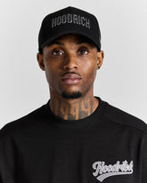 Man wearing a black 'Hoodrich' cap and sweatshirt against a gray background