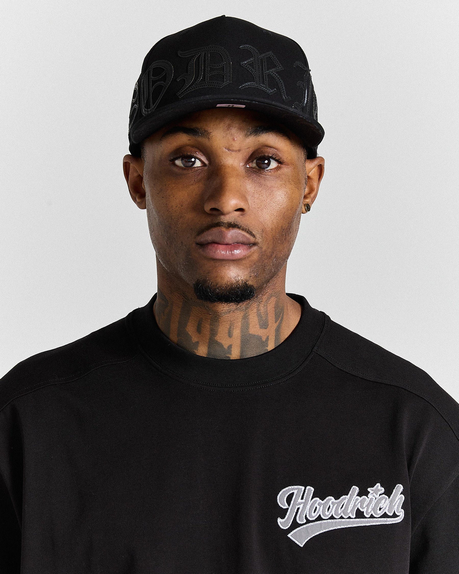Man wearing a black cap and black 'Hoodrich' sweatshirt on a gray background
