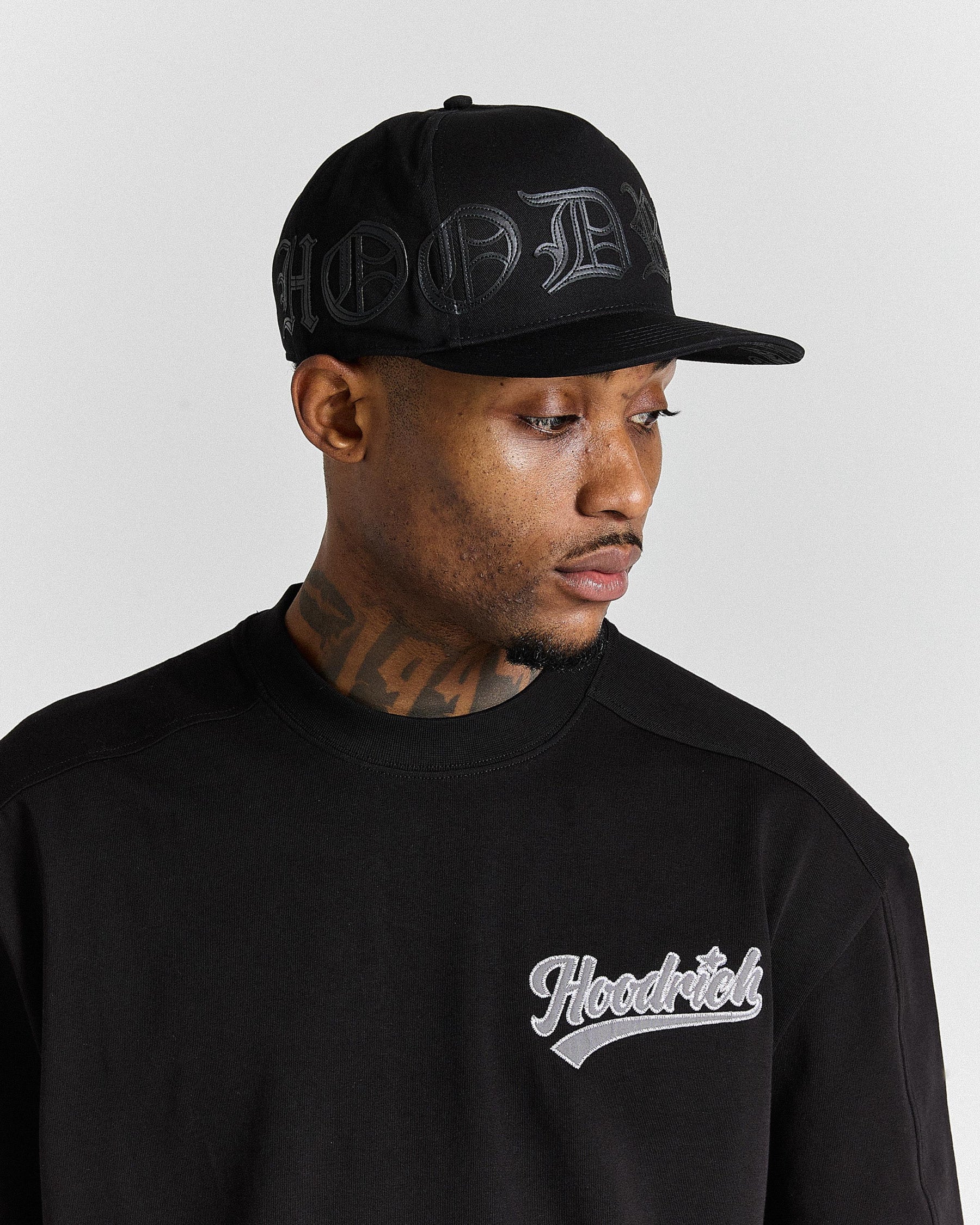 Man wearing a black cap and black sweatshirt with 'Hoodrich' logo on a gray background