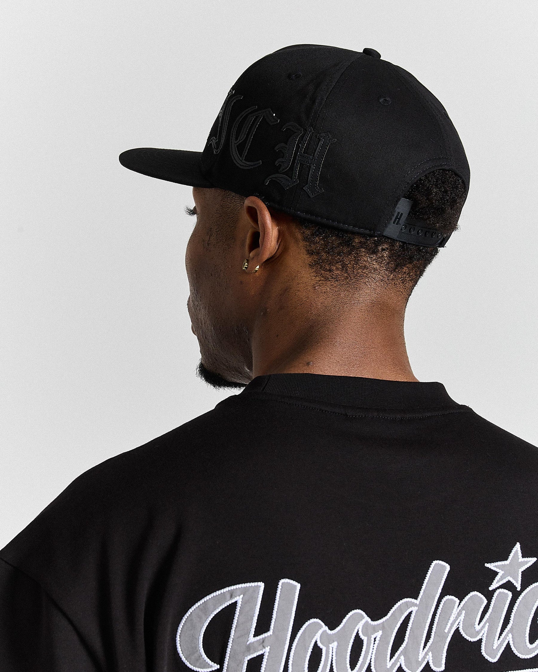 Person wearing a black cap and black 'Hoodrich' shirt with a star on a light gray background
