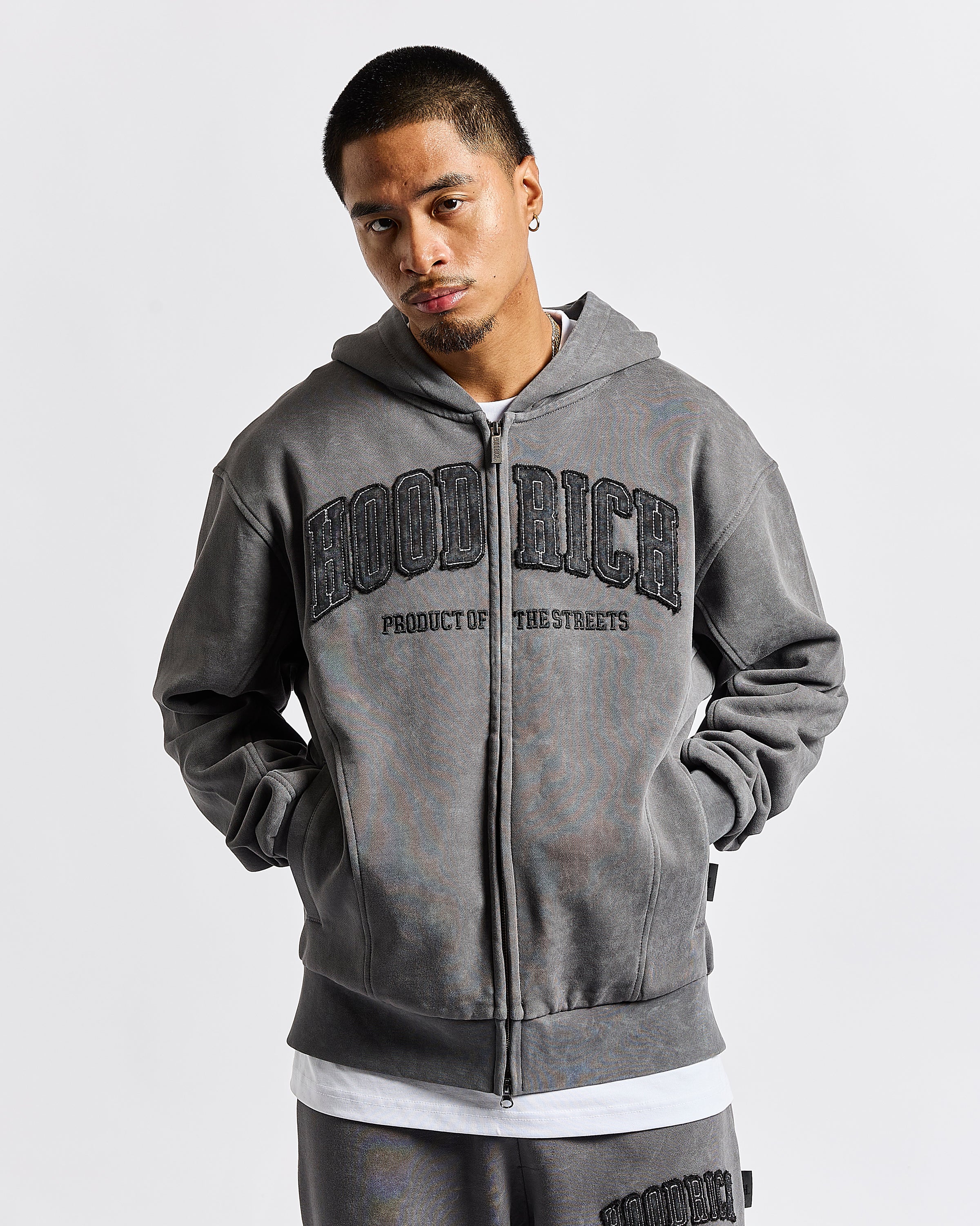 A man wearing a dark gray zip-up hoodie with 'HOOD RICH' printed across the chest, paired with matching gray sweatpants, against a light gray background.