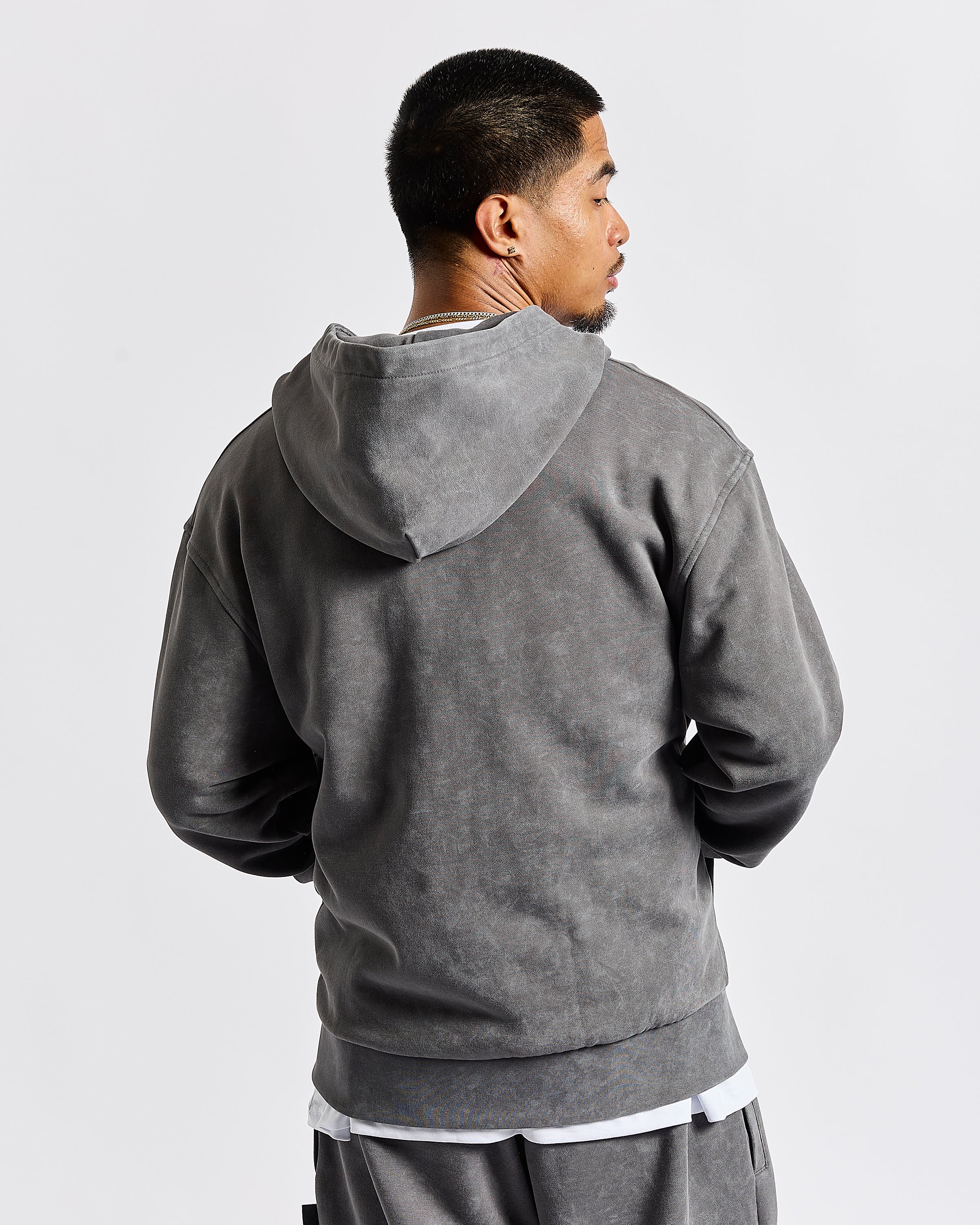 A man wearing a dark gray washed hooded sweatshirt, shown from the back and side profile against a light gray studio background.