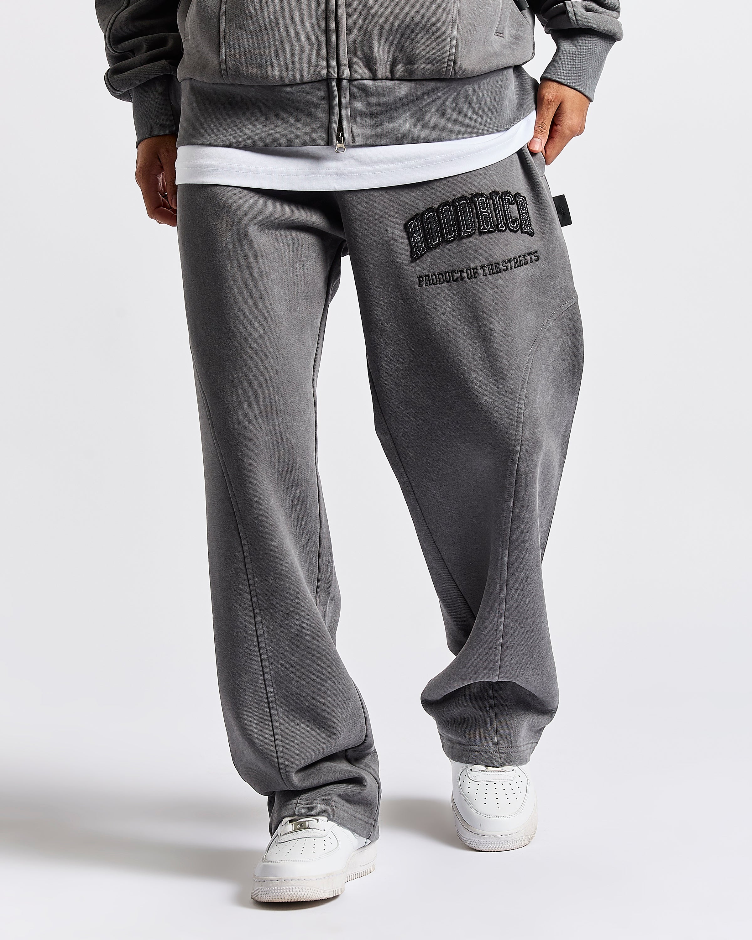 A person wearing gray washed sweatpants with black 'HOODRICH' and text on the left thigh, styled with a white undershirt and white sneakers against a light gray studio background.