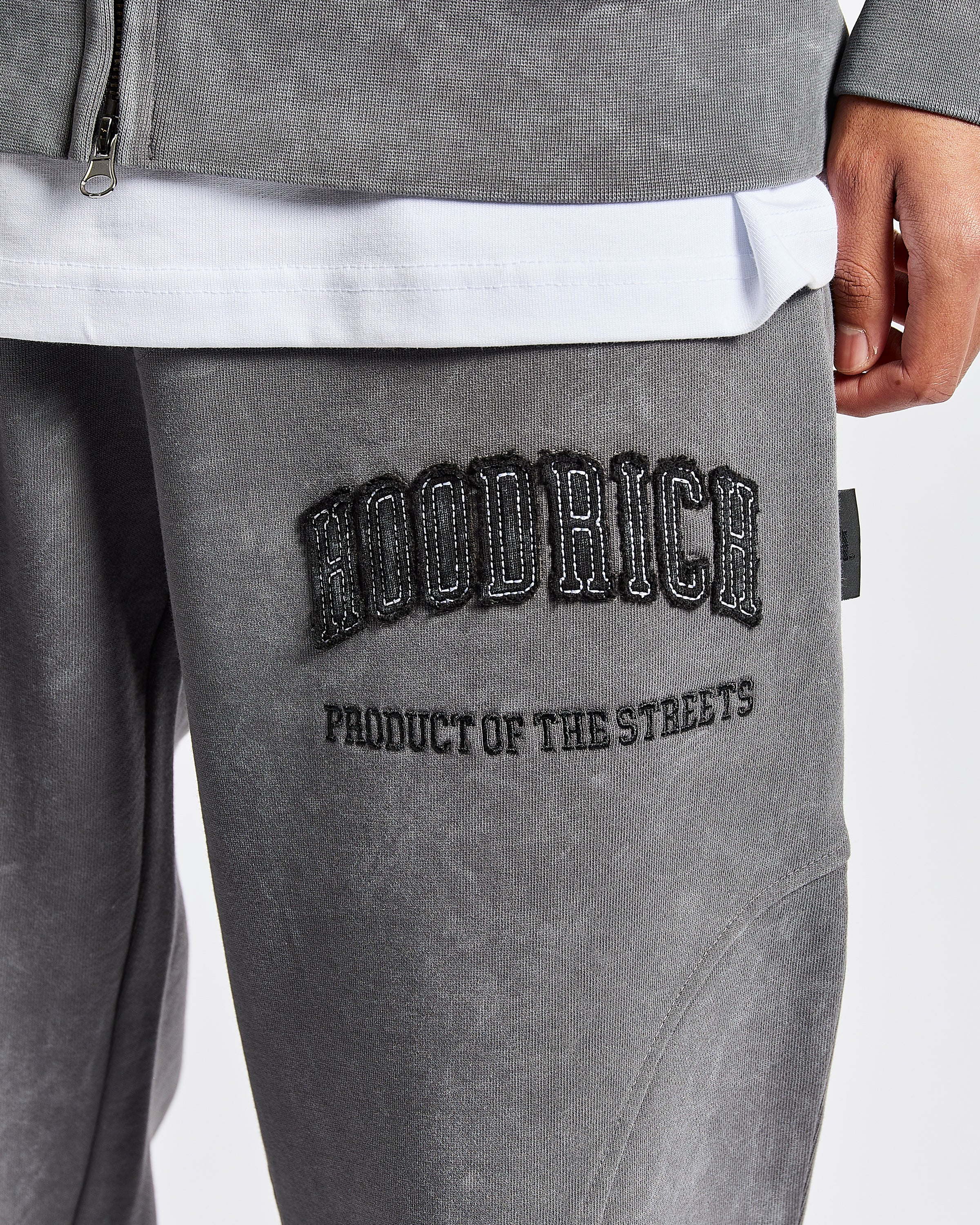 Close-up of a person wearing gray hooded sweatpants with black embroidered text reading 'HOODRICH' on the thigh, plus a white undershirt and gray zip jacket visible.
