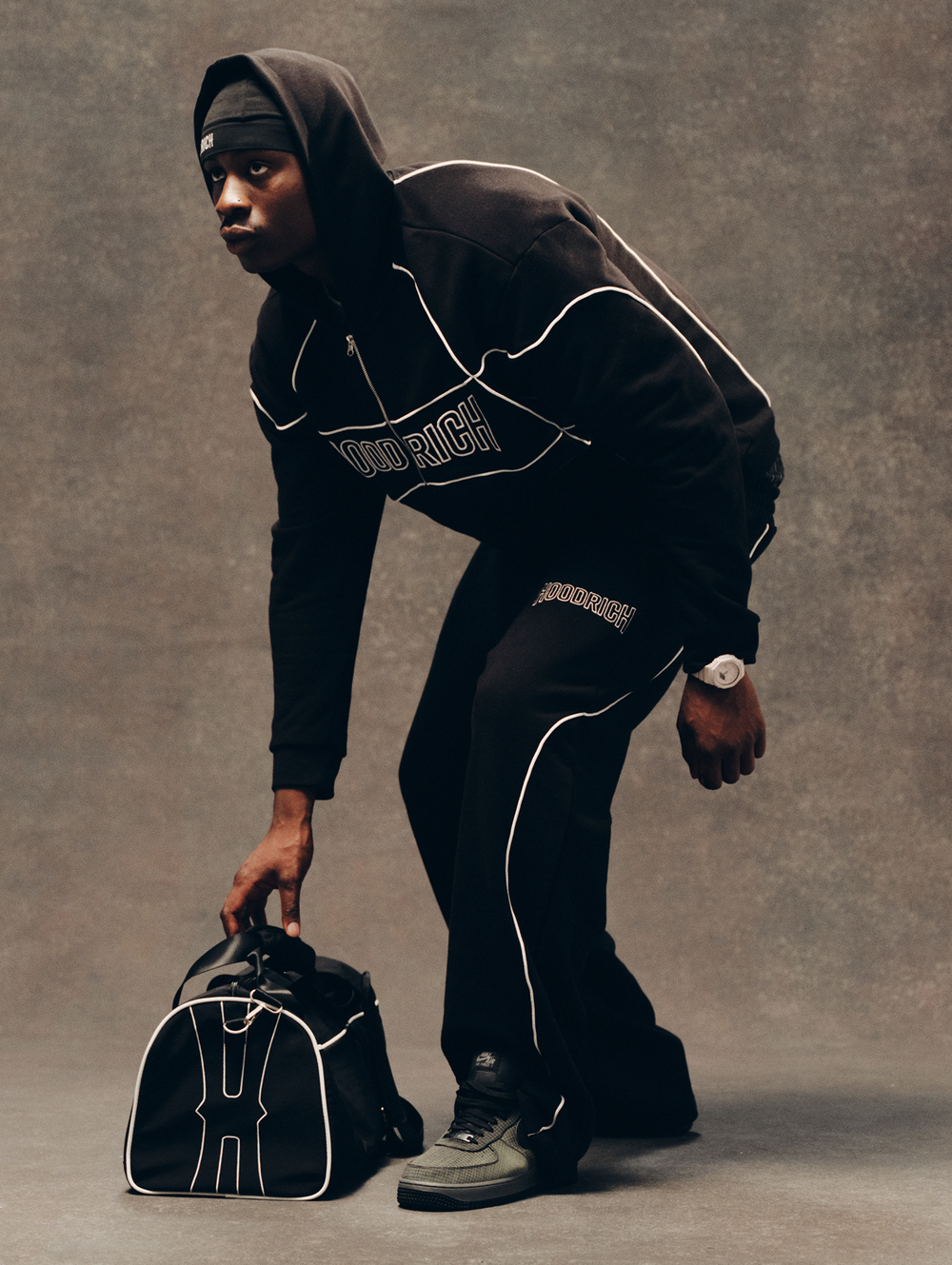 A man in a black hooded tracksuit with white piping and 'HOODRICH' text on the chest, crouching while picking up a black duffel bag with white piping against a textured gray studio backdrop.